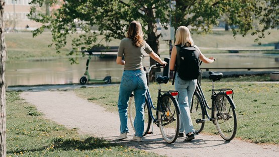 Två kvinnor går bredvid varandra på en grusväg och leder varsin cykel.