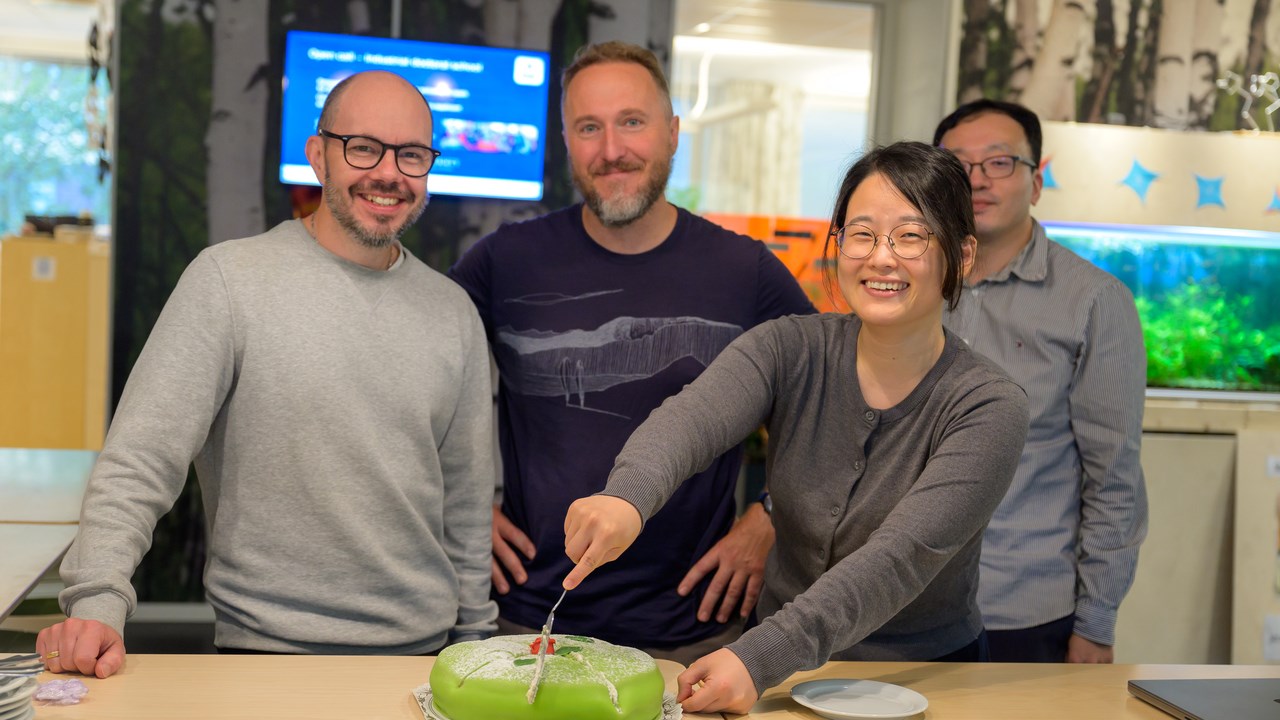 Ludvig Lizana, Eric Libby and Hye Jin Park smile while Hye Jin cuts a green cake. Sang Hoon Lee is in the background.