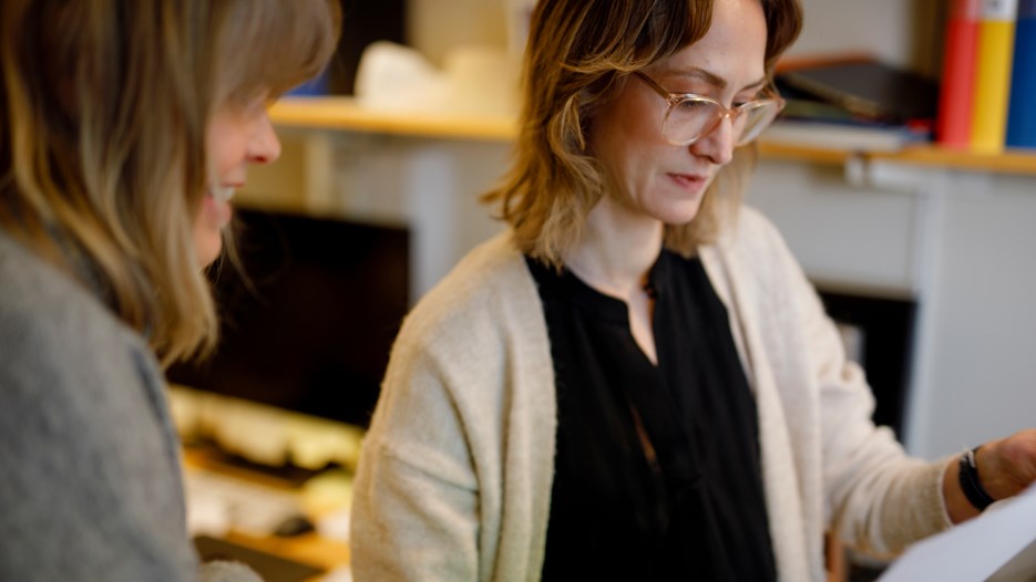 A close-up picture of Ulrika Nordström and Angelica Nordin, standing in an office looking at a sheet in a binder out of frame. 