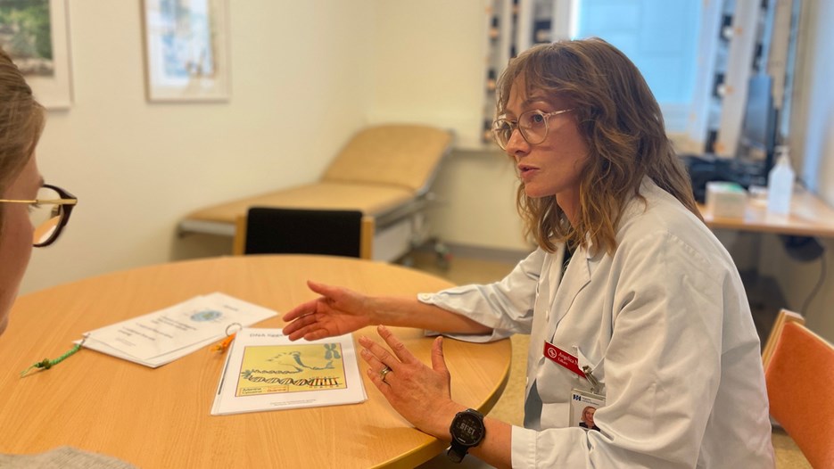 : In an examination room, Angelica Nordin is seen sitting at a round table talking to someone mostly out of frame. She is using her hands, explaining the contents of a couple of leaflets. In the background a bed and a desk with a computer and a bottle of hand sanitiser is seen. 