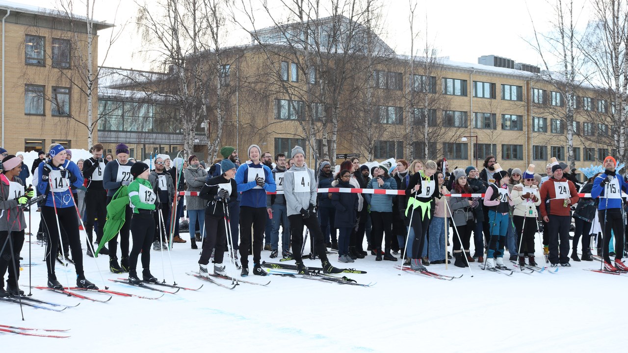 Several people on skis with numbers on their breast are waiting in front of  a white and red plastic band for their turn in the skiing relay. Behind of the plastic band, people cheering for the teams are standing.