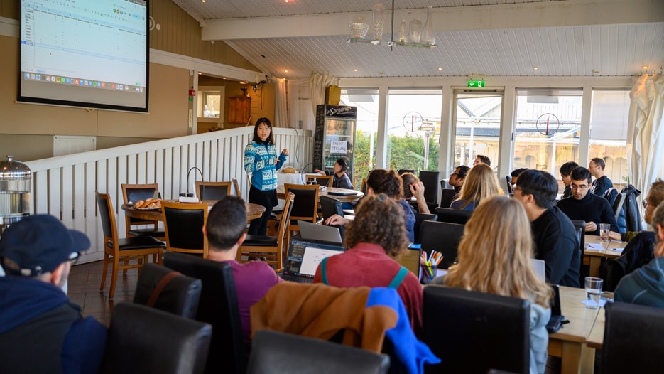 A group of people sitting around tables in a room listen to a woman giving a presentation