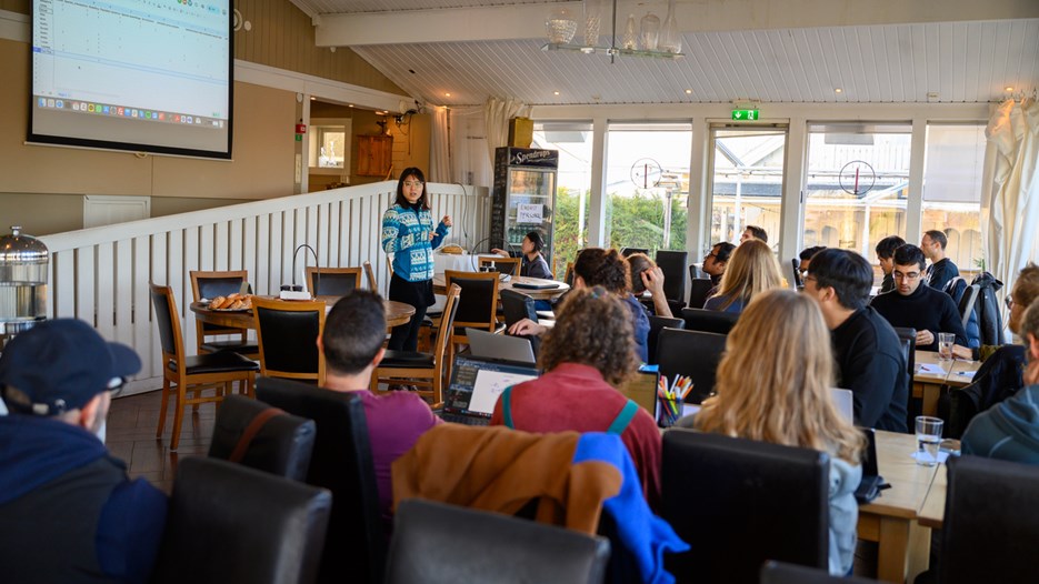 A group of people sitting around tables in a room listen to a woman giving a presentation