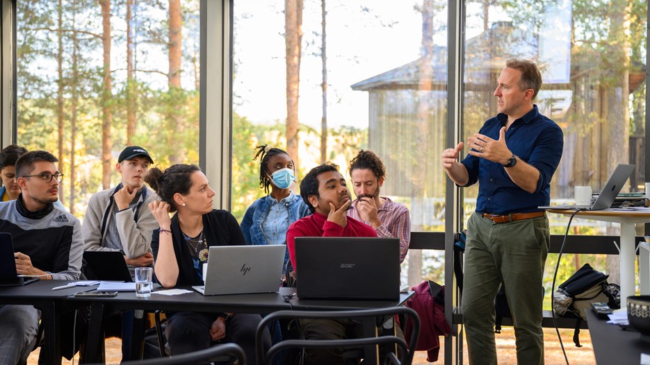 A group of students sitting at tables with laptops open listen to a man standing and speaking while gesturing with his arms. They are in a glass enclosed room, trees and another wooden house are visible outside. 