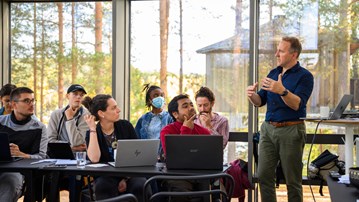 A group of students sitting at tables with laptops open listen to a man standing and speaking while gesturing with his arms. They are in a glass enclosed room, trees and another wooden house are visible outside.