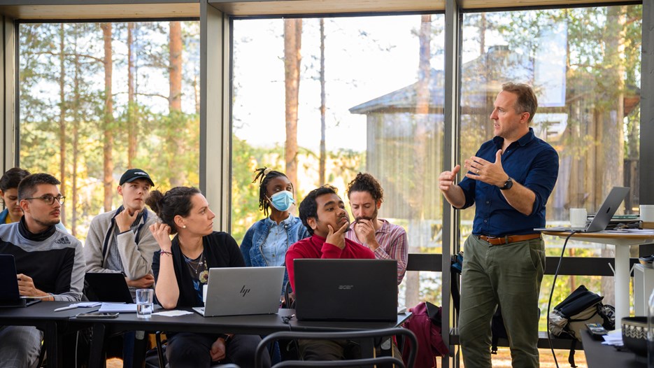 A group of students sitting at tables with laptops open listen to a man standing and speaking while gesturing with his arms. They are in a glass enclosed room, trees and another wooden house are visible outside. 