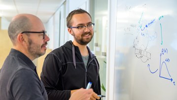 Two men look at drawings on a whiteboard