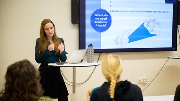 Verena Kohler ger en pitch medan hon står framför en skärm som visar en bild där det står "var behöver vi akademiska vänner".Verena Kohler gives a pitch while she stands in front of a screen that shows a slide that says "where do we need academic friends".