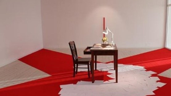 Ett konstverk av ett skrivbord och stol med papper utspridda på en röd matta i form av ett kryss.
