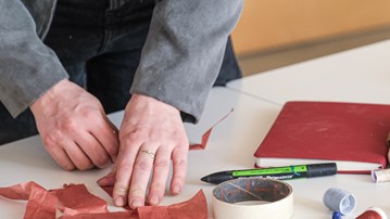 Händer som arbetar med silkespapper på ett bord med broderigard och pennor.