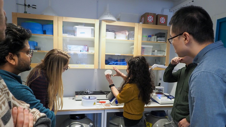 Students standing around the teacher in a lab, the teacher shows different tools to be used. Some students take notes.