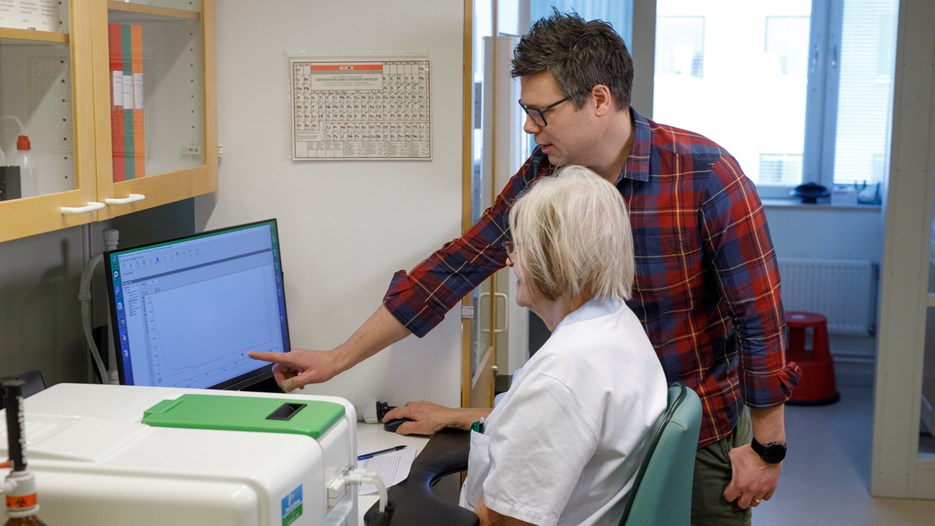 Karin Hjertkvist is sitting by a computer on a lab bench, looking at the results of an enzymatic measurement. Standing next to her is Per Zetterström, commenting on something he is pointing at on the screen. 