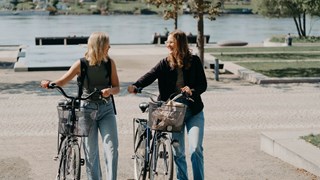 Två kvinnor går bredvid varandra och styr varsin cykel.