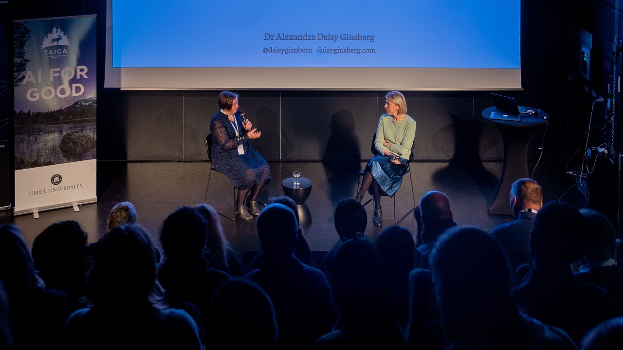 two people having a conversation in front of an audience in a dark room