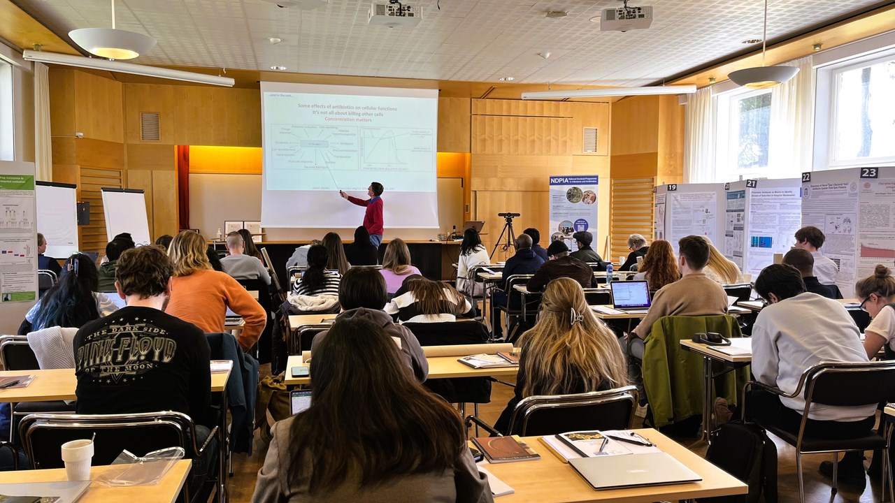 A Professor is giving a lecture and pointing on the projector screen with a pointing stick. The lecture hall is full of students and the photo is taken from the back of the room.