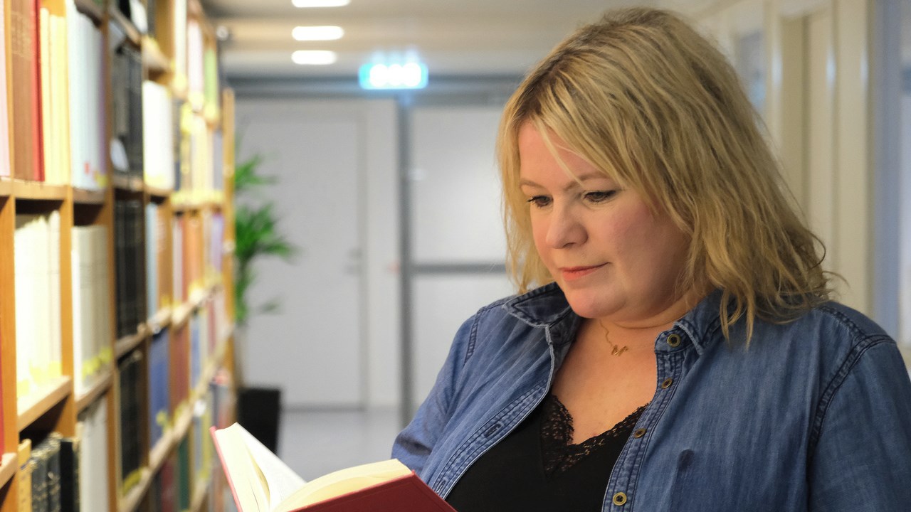 Maria Forsman står vid bokhylla med en bok i händerna.