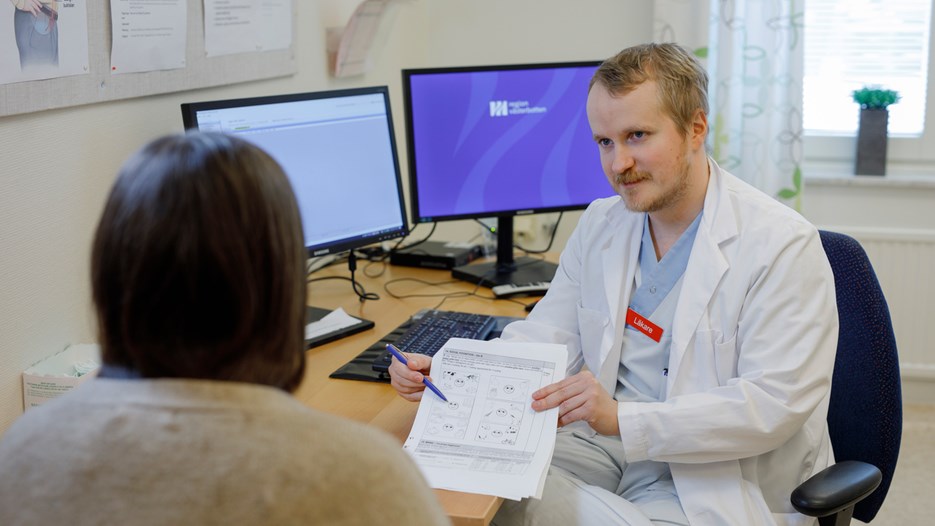 A person is sitting in an examination room with their back against the camera, looking at a form that Ivar Winroth is holding up. In the background, a desk with computer screens can be seen.