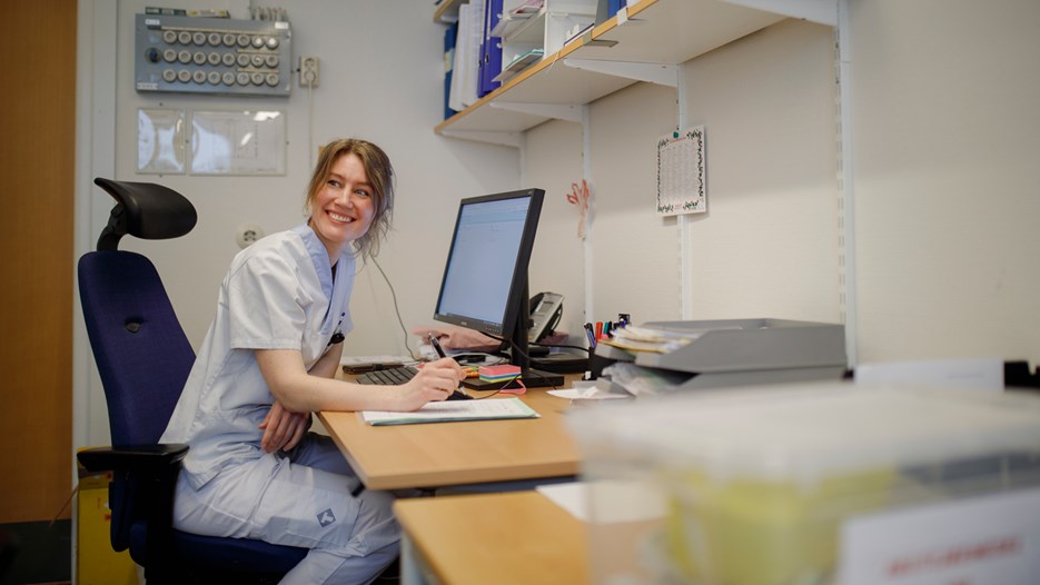 Tina Björn is seen sitting by her computer, smiling and looking over her shoulder at a collegue out of frame.