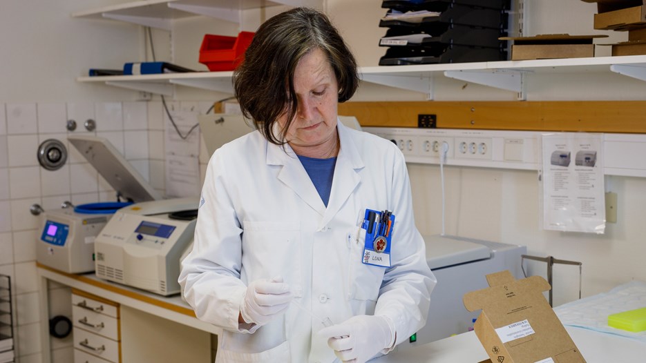 : Lena Bylund is standing next to a lab bench, pipetting samples from a test tube. On the bench is an opened cardboard box that has arrived at the clinic. In the background, several bench top centrifuges can be seen.