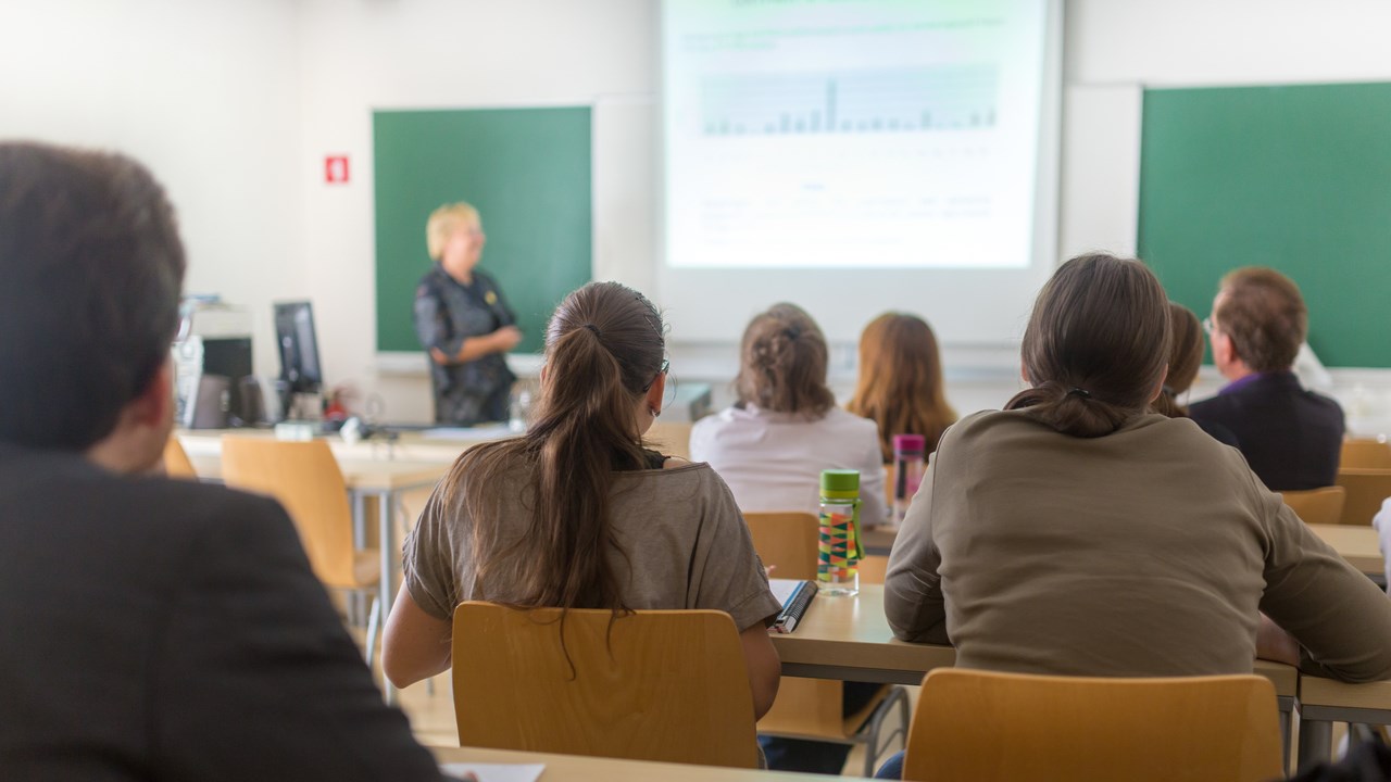 Föreläsningssal på universitet med studenter i förgrunden och en lärare vid tavlan.