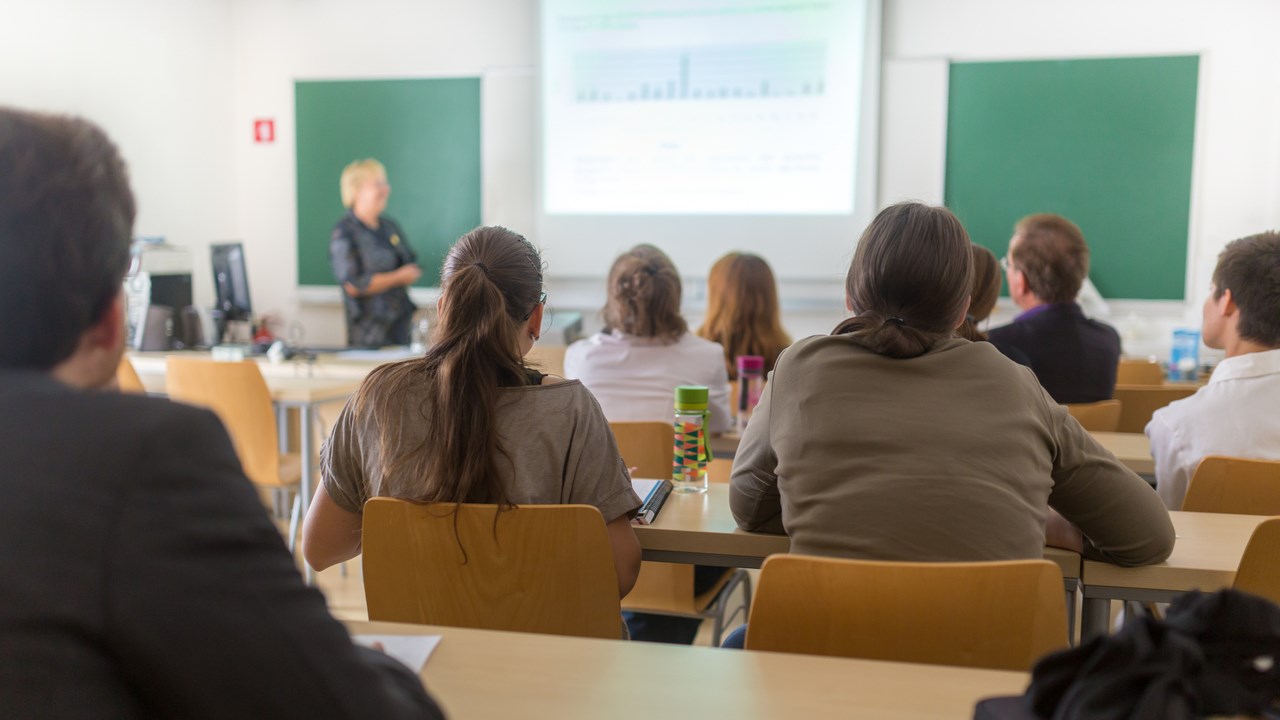 Föreläsningssal på universitet med studenter i förgrunden och en lärare vid tavlan.