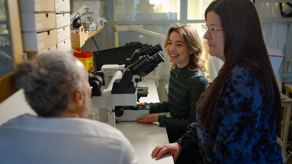 A smiling Isabelle Sigfridsson is sitting by a microscope discussing her findings with Matthew Marklund and Sara Rimpi, who are using the microscope’s two extra sets of oculars.