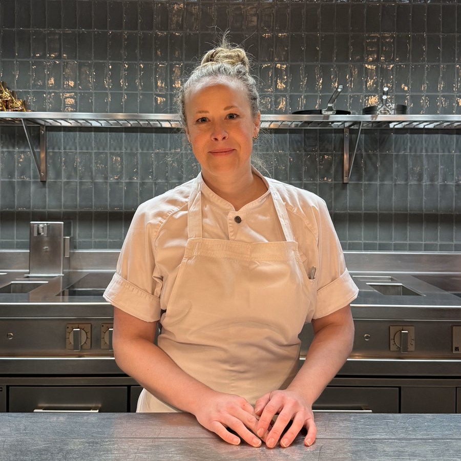 Emma Wixner, står i ett restaurangkök med kökskläder i vitt och grönt kakel i bakgrunden. Samt spis och hyllor i aluminium syns i bakgrunden.