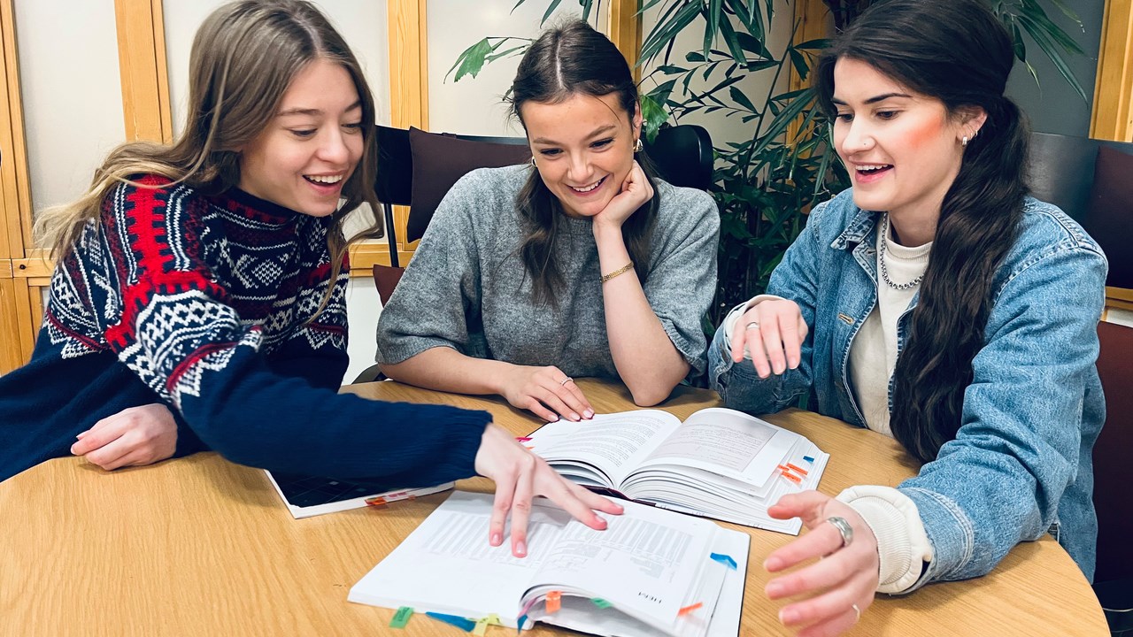 Bild på studenterna Elsa Nydén, Olivia Lundberg och Eira Bergström von Euler som bläddrar i läroböcker, sittande vid ett runt bord.