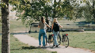 Två kvinnor går bredvid varandra på en grusväg och leder varsin cykel. 