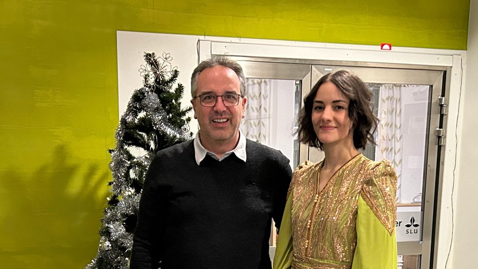 Nabila El Arbi in a green dress stands together with her supervisor Markus Schmid in front of a Christmas tree and a green-yellowish wall