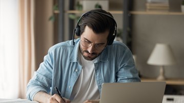 En man sitter vid ett bord med laptop och headset, böcker och antecknar i ett block.