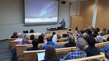 Hörsal fylld med människor som sittandes kollar mot talaren. Talaren visar upp en bild på norrsken på en stor projektorskärm.