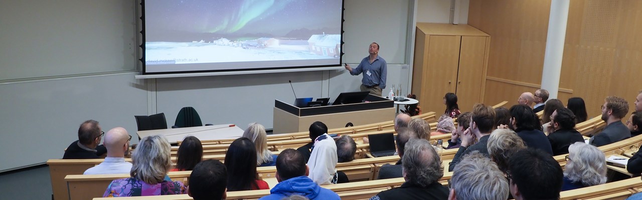 Hörsal fylld med människor som sittandes kollar mot talaren. Talaren visar upp en bild på norrsken på en stor projektorskärm.