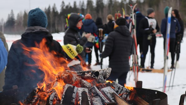 Group of people with and without skis standing outside. In the front, the wood is burning in the outdoor firepit