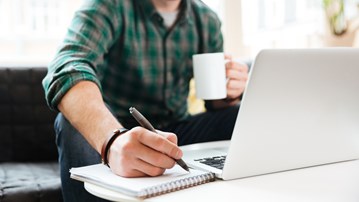 Person sitter i en soffa och arbetar. Har dator, kaffekopp och block där han skriver.