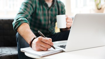 Person sitter i en soffa och arbetar. Har dator, kaffekopp och block där han skriver.