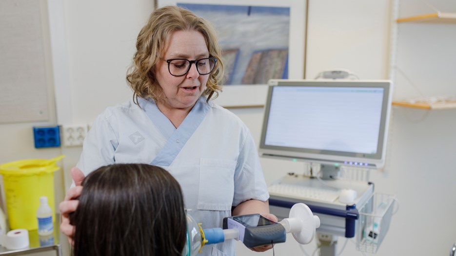 I ett undersökningsrum med med medicinsk utrustning står forskningssjuksköterskan och studiekoordinatorn och håller en spirometer mot en persons mun. Andra handen håller hon om personens bakhuvud och hon tittar på spirometerns display.