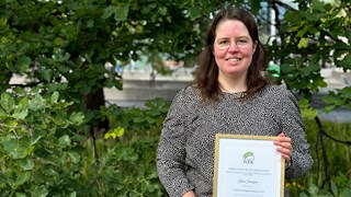 Anna Jonsson, universitetslektor i datavetenskap, har långt hår och glasögon. Hon står i en grönskande utemiljö, och håller i sitt diplom.