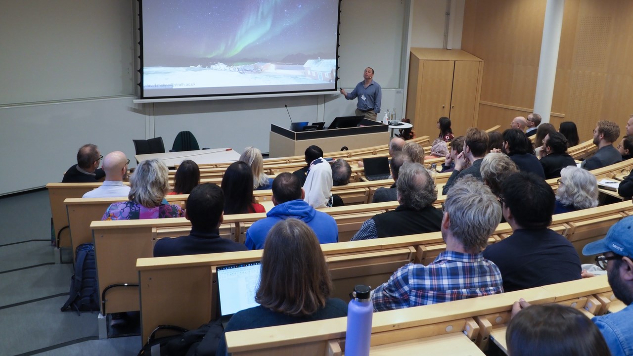 Hörsal fylld med människor som sittandes kollar mot talaren. Talaren visar upp en bild på norrsken på en stor projektorskärm.