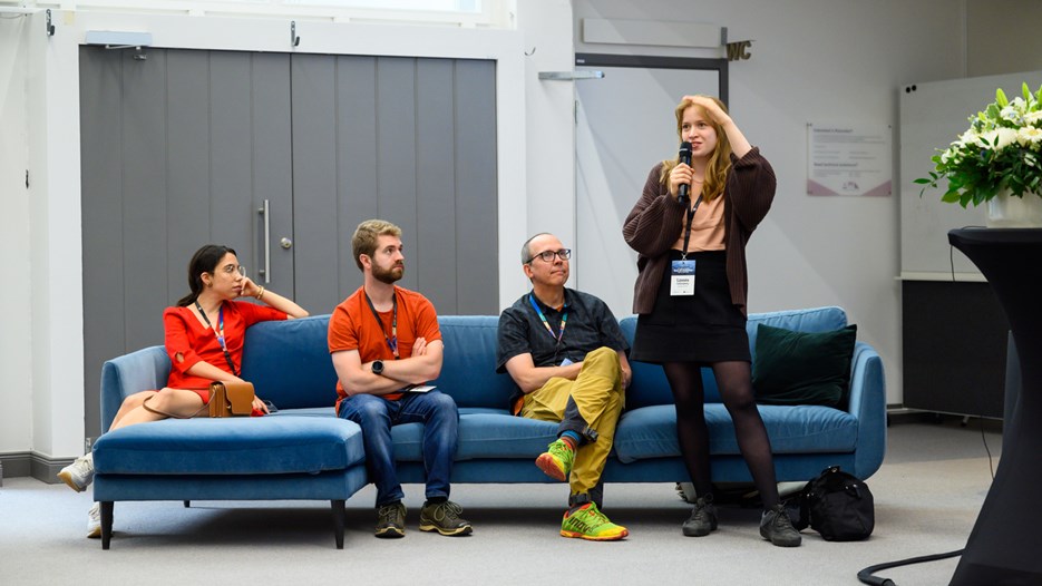 Three people sit on a blue couch and listen to Linnea standing and speaking while holding a microphone.