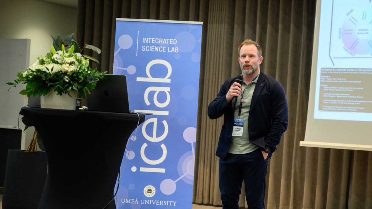 Mattias Marklund stands holding a microphone, next to a large screen and a blue roll-up with the word IceLab displayed.