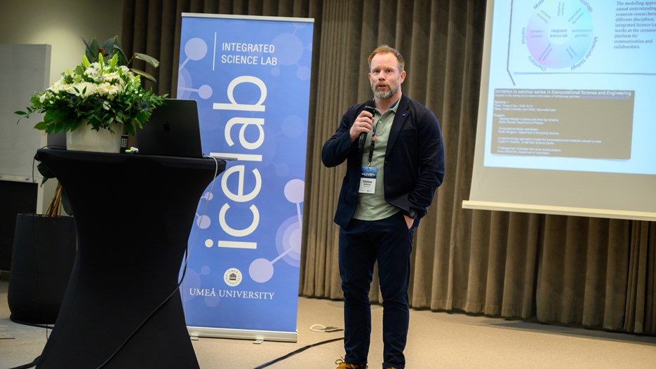 Mattias Marklund står och håller i en mikrofon, bredvid en stor skärm och en blå roll-up med ordet IceLab.