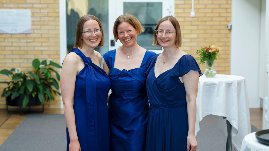 Tre systrar i kornblå klänningar står uppställda framför kameran. 