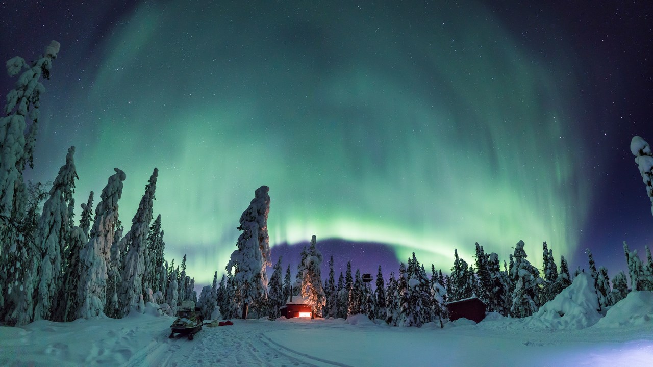 Upplyst stuga på vintern med ett stort grönt norrsken på den svarta himlen. Stugan står vid skogsbryn.