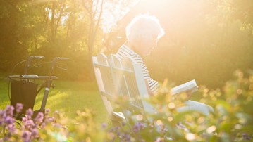 Äldre kvinna sitter i trädgård och läser bok i kvällsljus.
