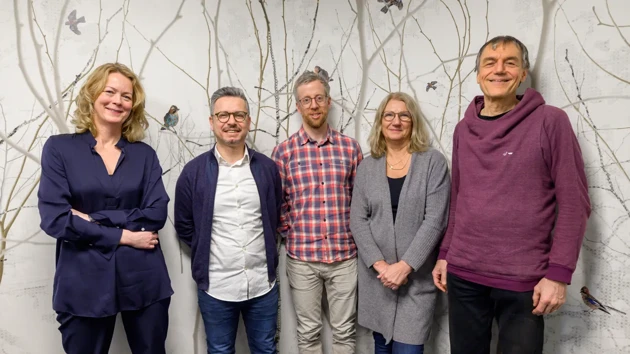 Five people standing in front of a wallpaper of a forest with birds