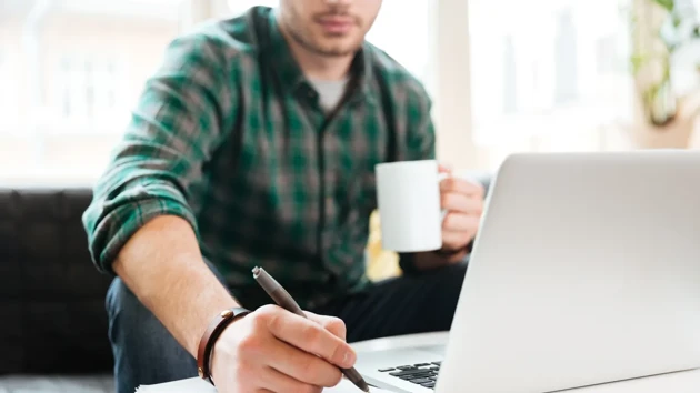 Man som sitter framför en dator med en kaffekopp och skriver med en penna.