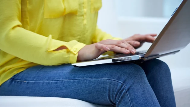 Person with a bright yellow shirt sitting on a sofa typing on a laptop.