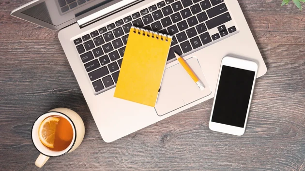 A computer, cup of tea, a notebook and a phone on a desk.