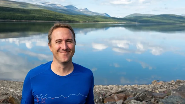 A man in a blue shirt in front of a lake reflecting the sky